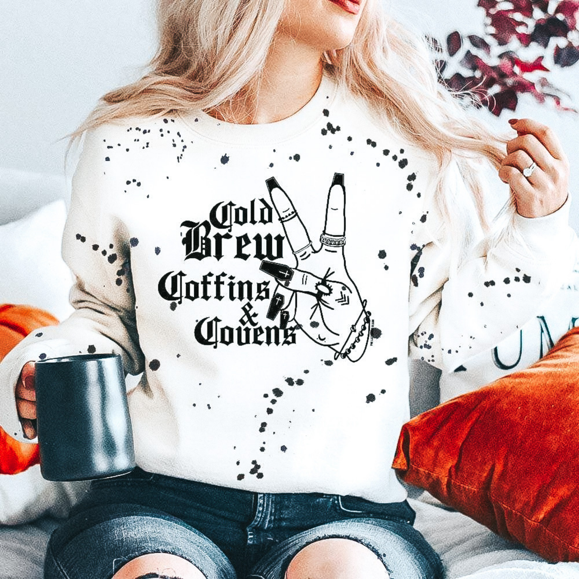 model is wearing a white, splatter paint sweatshirt which reads "cold brew, coffins, and covens" with a hand doing a peace sign.  Model is sitting in front of a fall background and has tee paired with black, ripped jeans. 