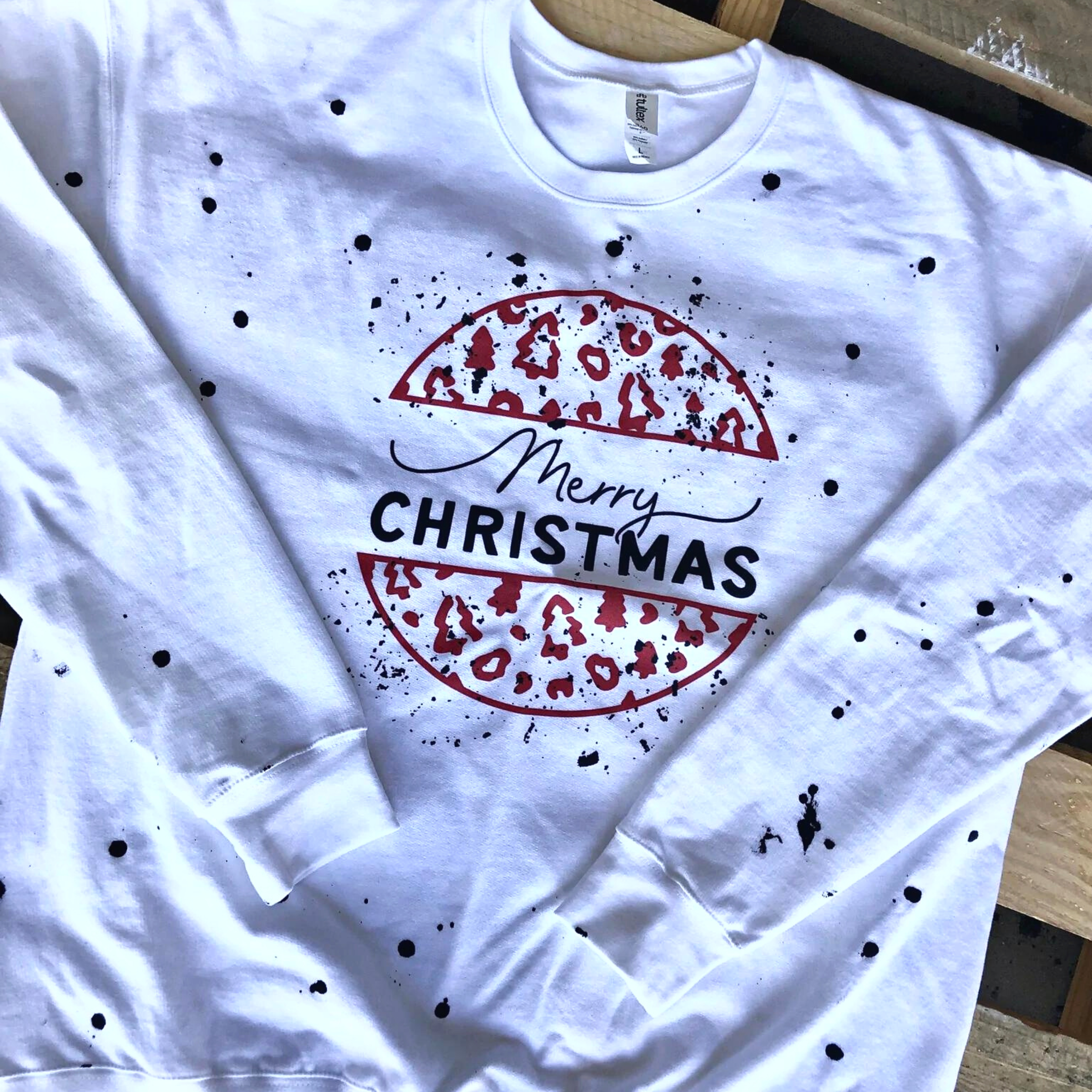 This white pullover includes a crew neckline, long sleeves, and a multi print Christmas tree collage graphic with black paint splatters all over. It is shown in this picture as a flat lay. 
