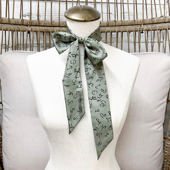 A sage green silk scarf with a black abstract pattern, tied in a bow and draped on a white fabric mannequin torso.
