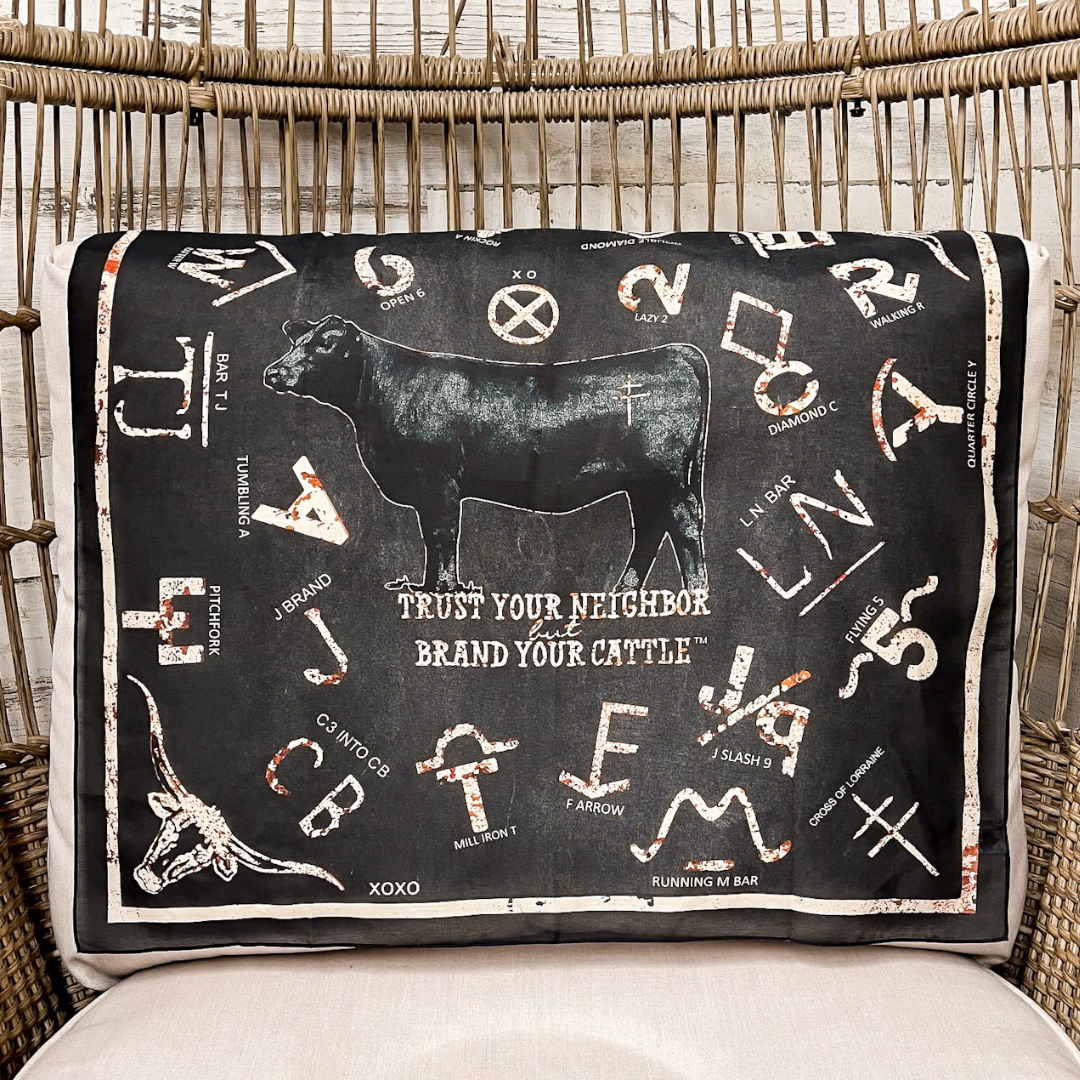 A scarf with a black background and white western-style graphics, including a cow silhouette, zodiac symbols, and the phrase 'TRUST YOUR NEIGHBOR BRAND YOUR CATTLE,' draped over a woven rattan chair.