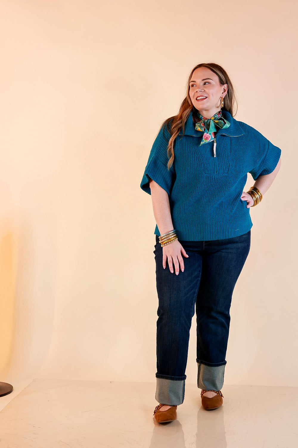 Model wearing a teal ribbed half zip sweater with a floral scarf, cuffed dark wash jeans and brown flats.