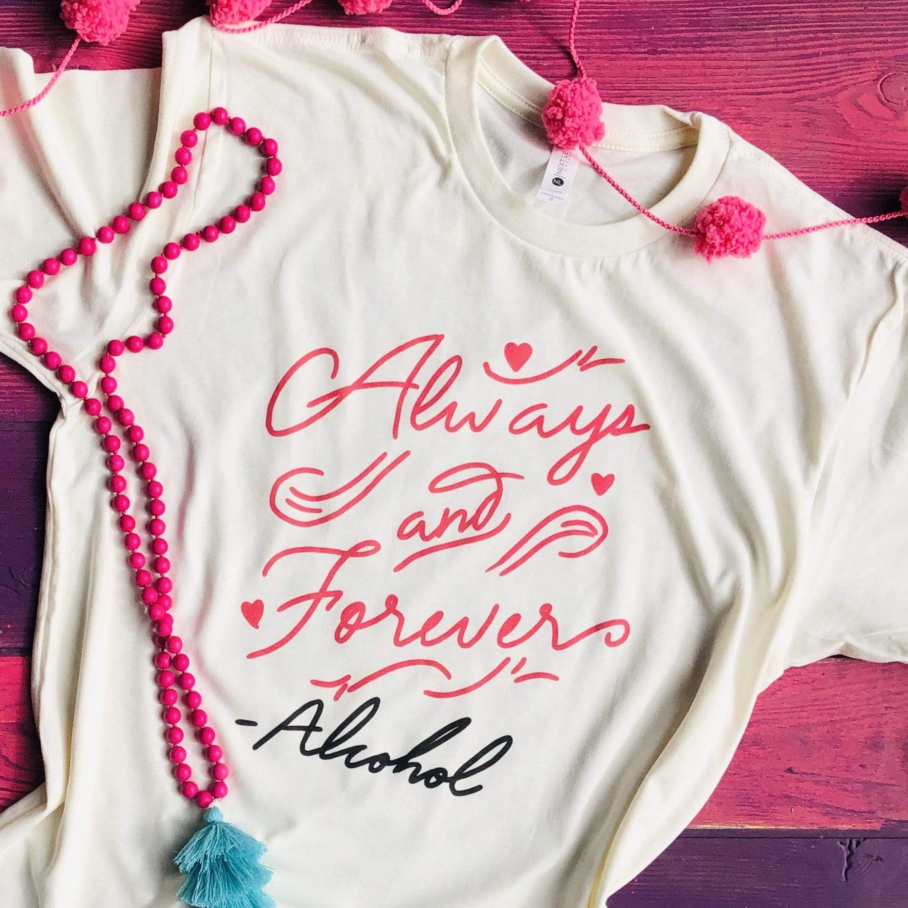 An ivory tee shirt laid on a purple wooden background, pictured with pink beads and pink puff balls. This tee shirt has a cursive graphic that says "Always and Forever" - Alcohol.
