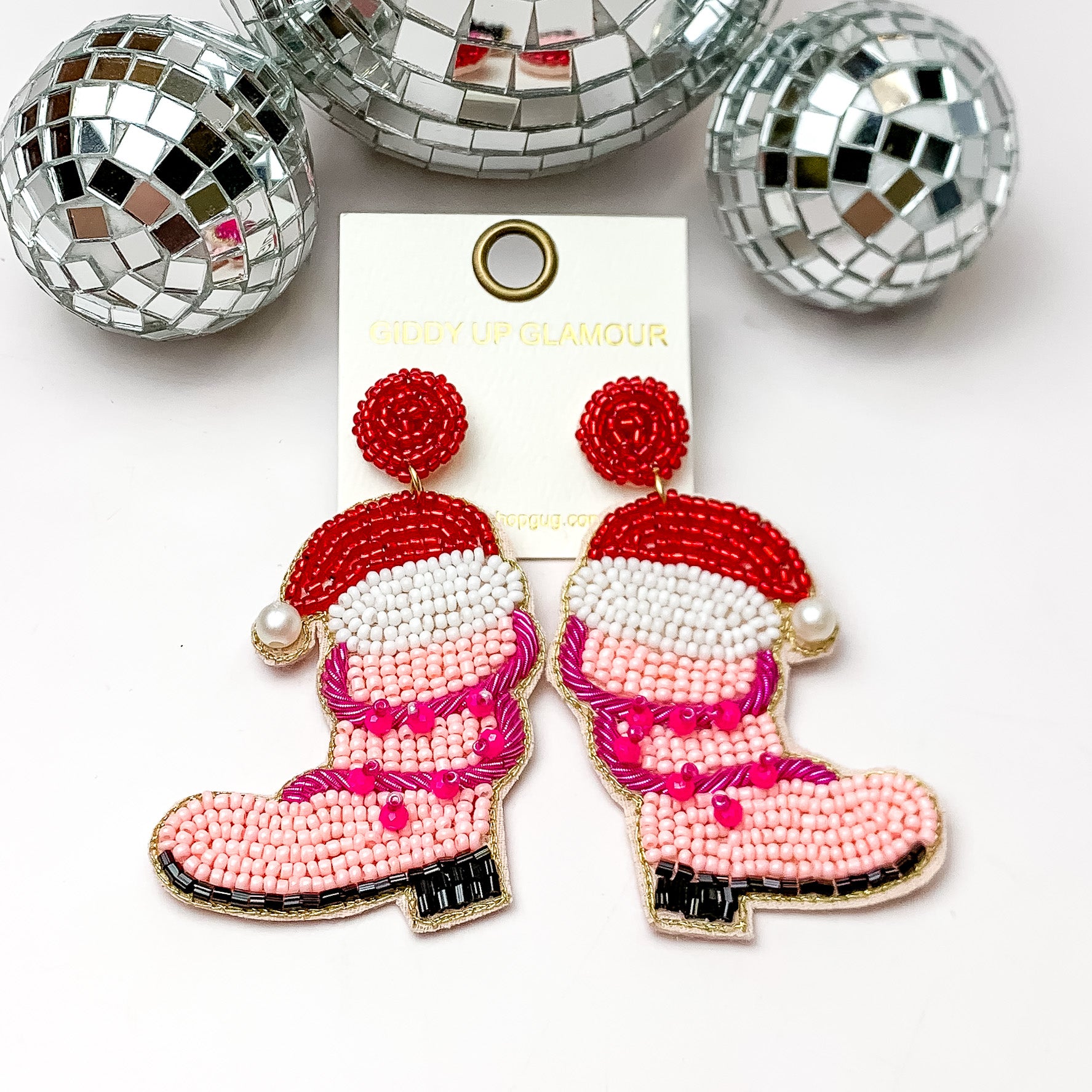 Pink beaded cowboy boot earrings with a red sant hat and fuchsia pink lights wrapped around them. These earrings are pictured on a white background with disco balls at the top. 