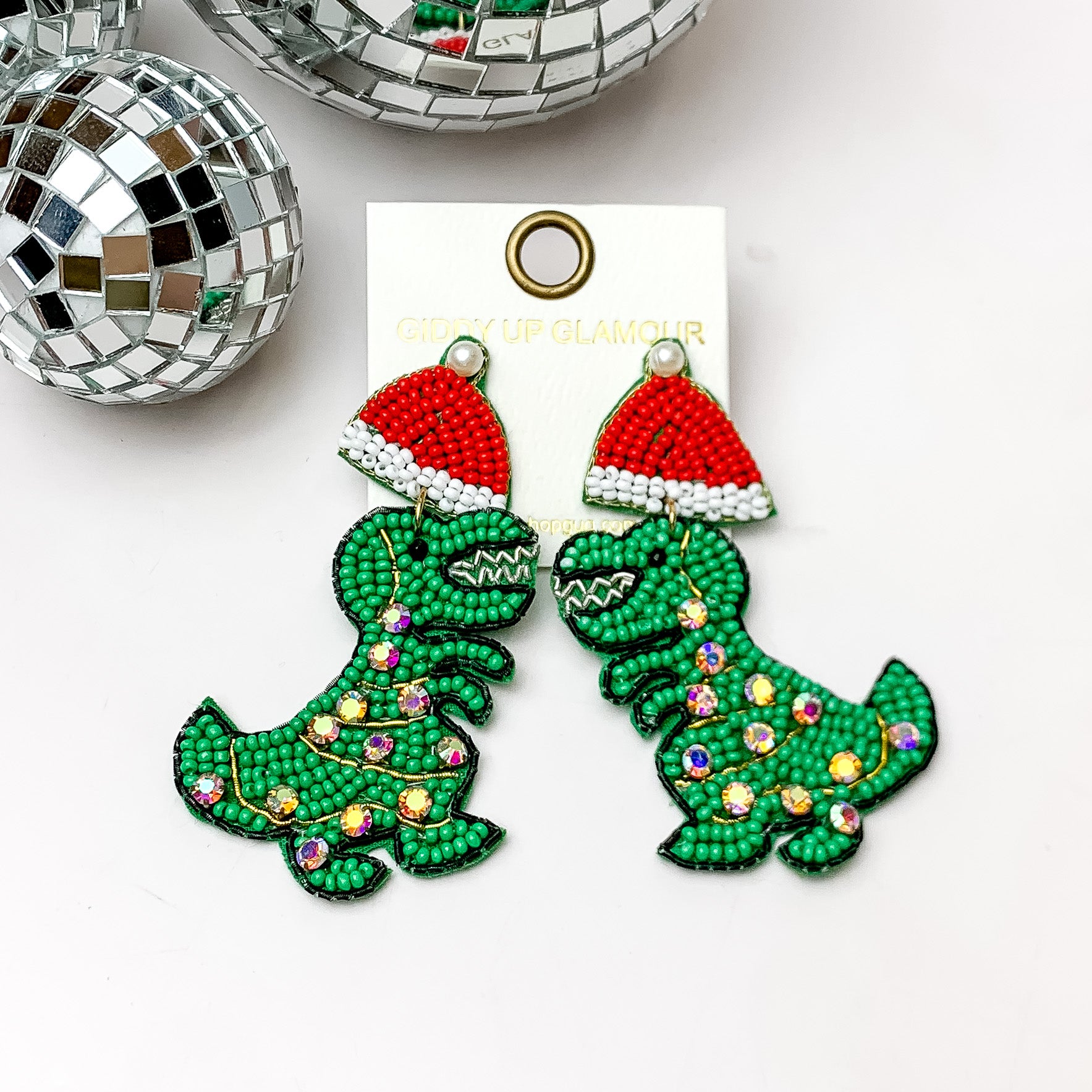 Green beaded trex earrings with red santa hat. These earrings are pictured on a white background with disco balls at the top. 