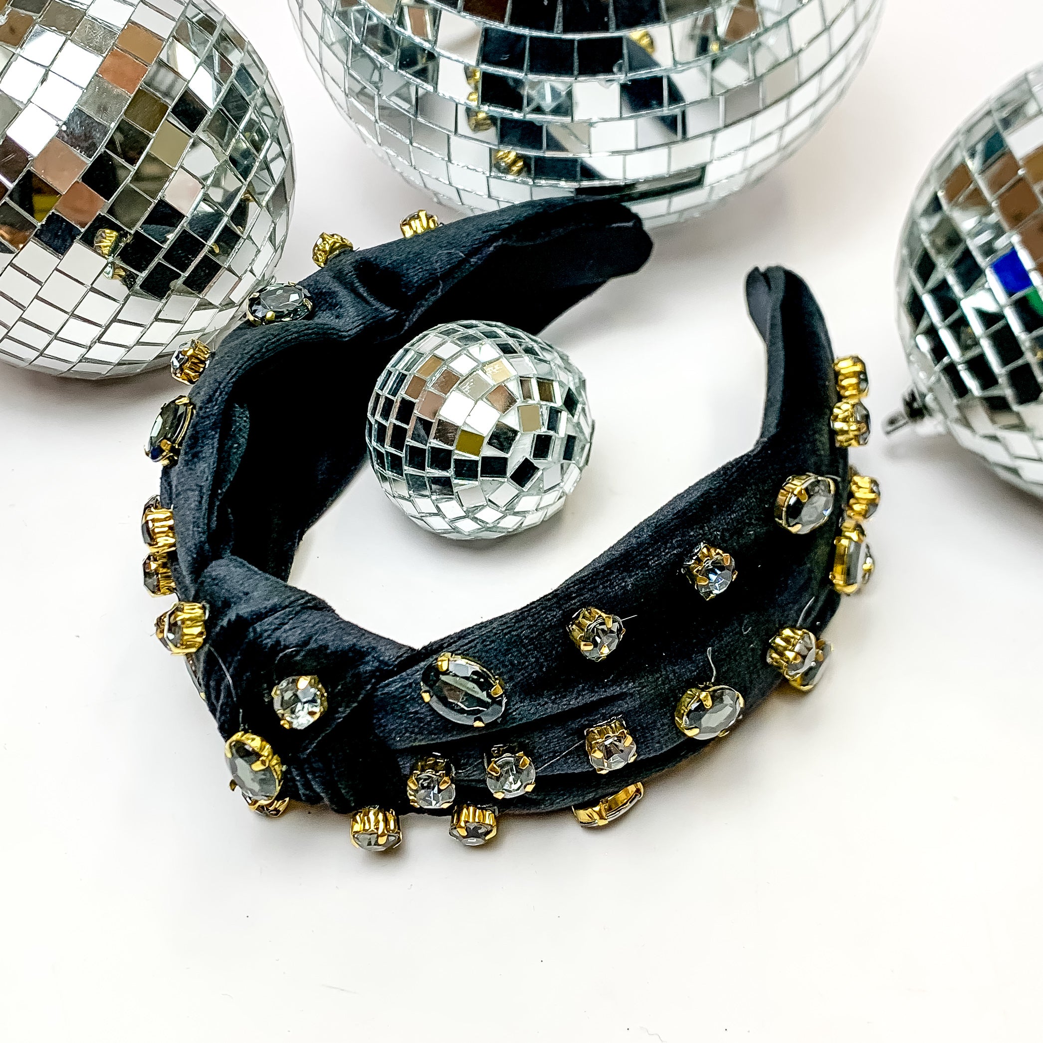 Crystal Detailed Velvet Knot Headband in Black. This headband is pictured on a white background and is surrounded by disco balls.