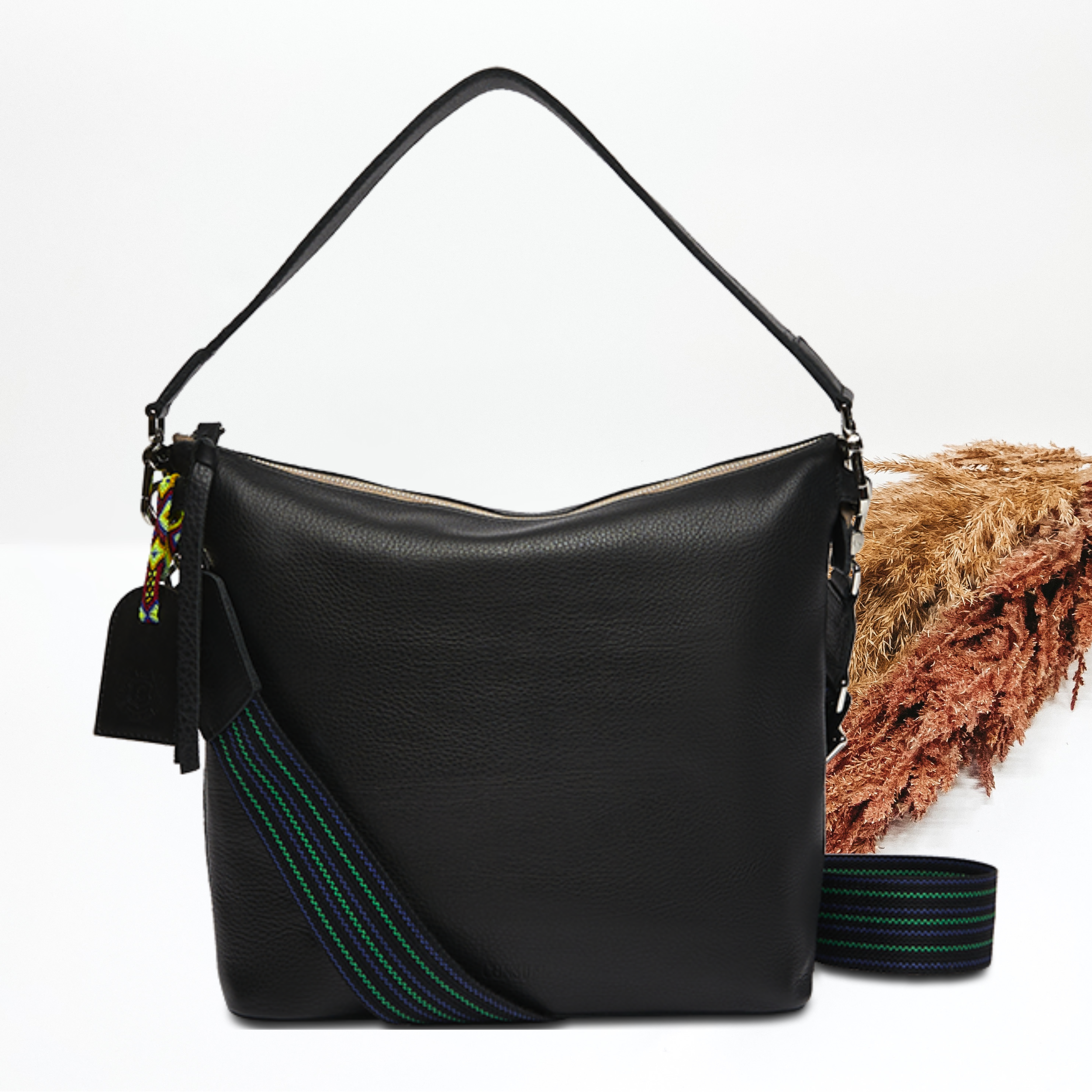 Pictured on a white background is black leather hobo bag with a black shoulder strap. This bag also includes a striped crossbody strap., black tassel on the zipper and a black charm.