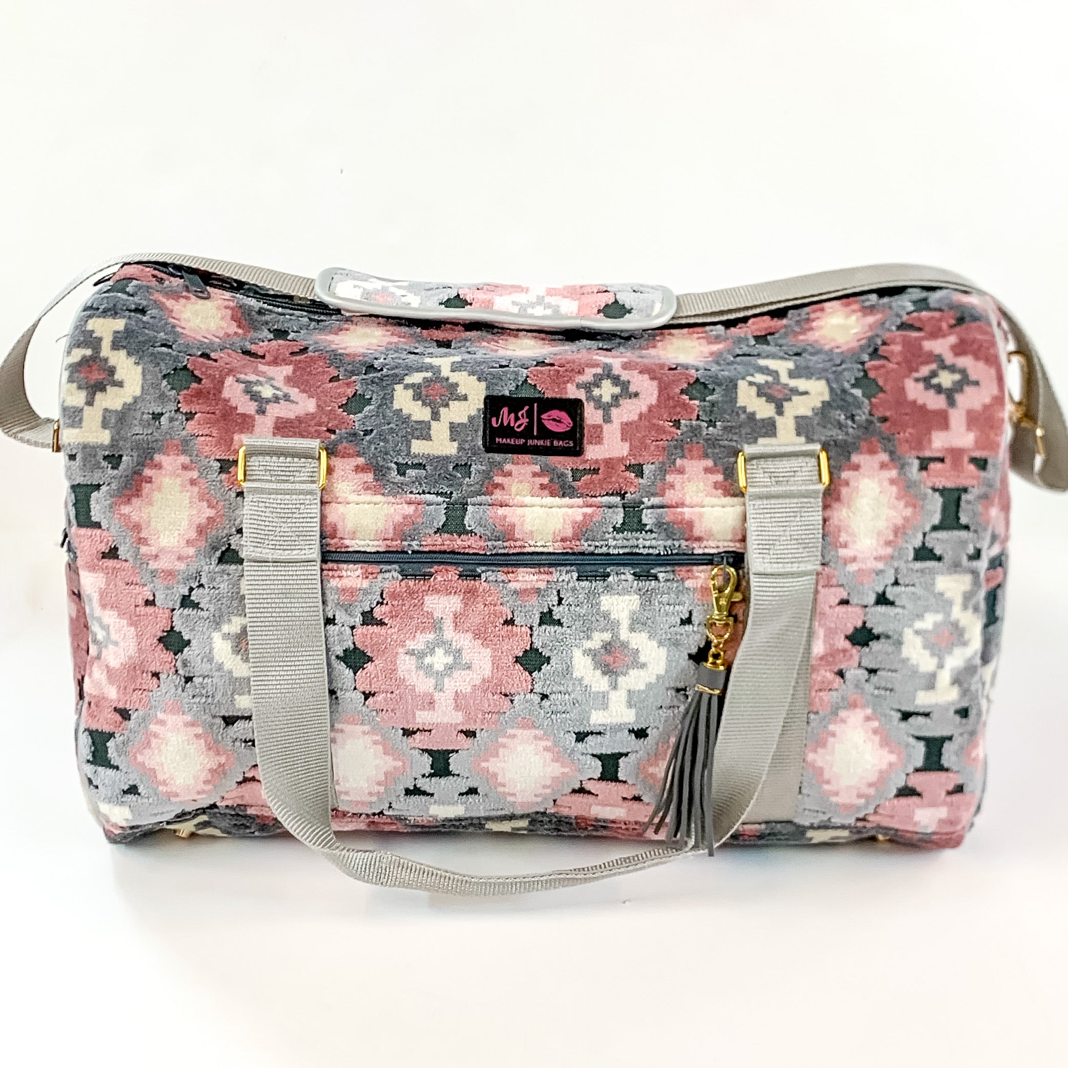 Pictured on a white background is a duffel bag with grey straps in a blush aztec print. This bag includes a zipper tassel, two short grey straps, and a grey tan strap.