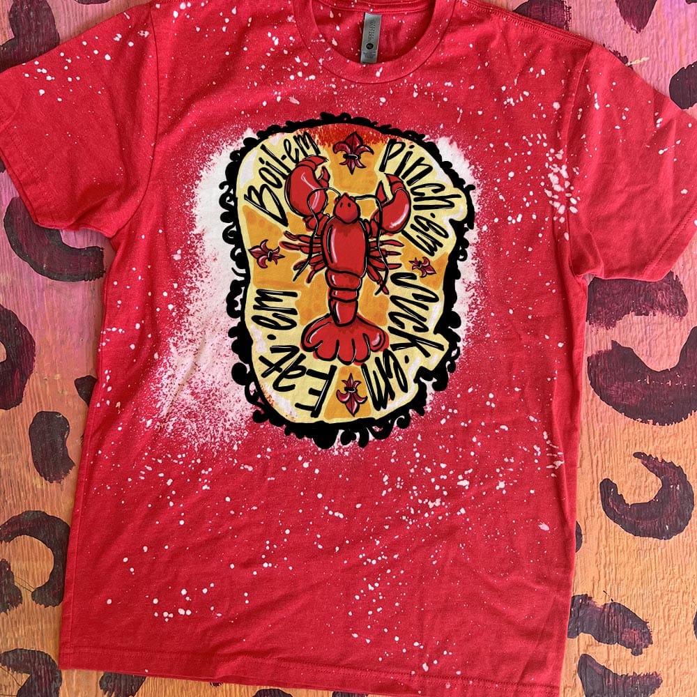 A red short sleeve shirt with a bleach splatter design and a large bleach spot in the center featuring a graphic of a red crawfish with an asymmetric orange circle behind it. The words "Boil-em, pinch-em, suck-em, eat-em" circle the crawfish and a black border can be found around the orange background.