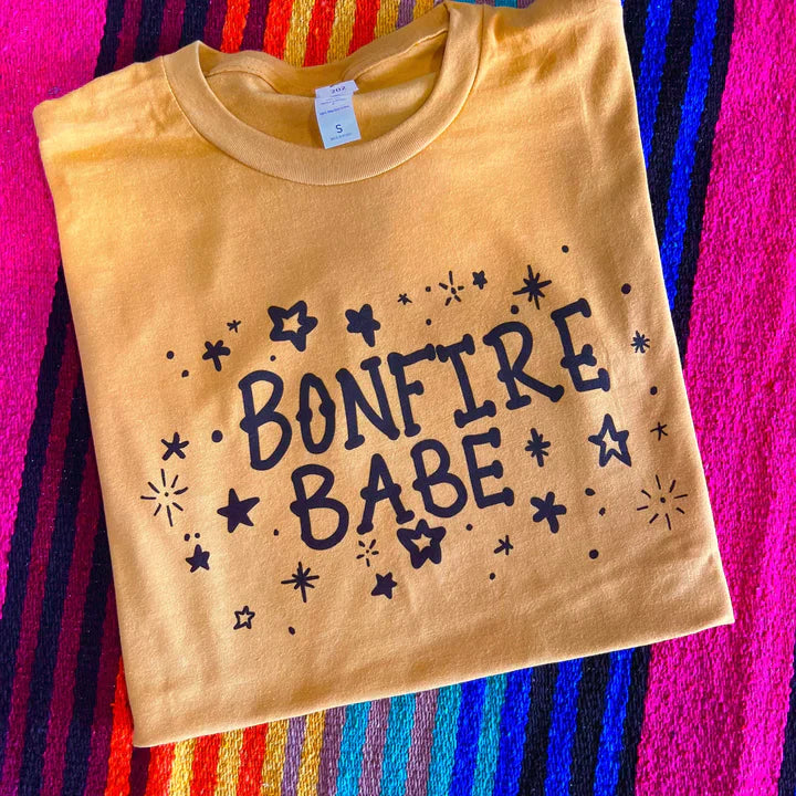 This mustard yellow tee includes a crew neckline, short sleeves, and a cute graphic of stars surrounding the words "BONFIRE BABE" in a fun, black font. This tee is pictured here as a folded flatlay on a colorful serape blanket. 
