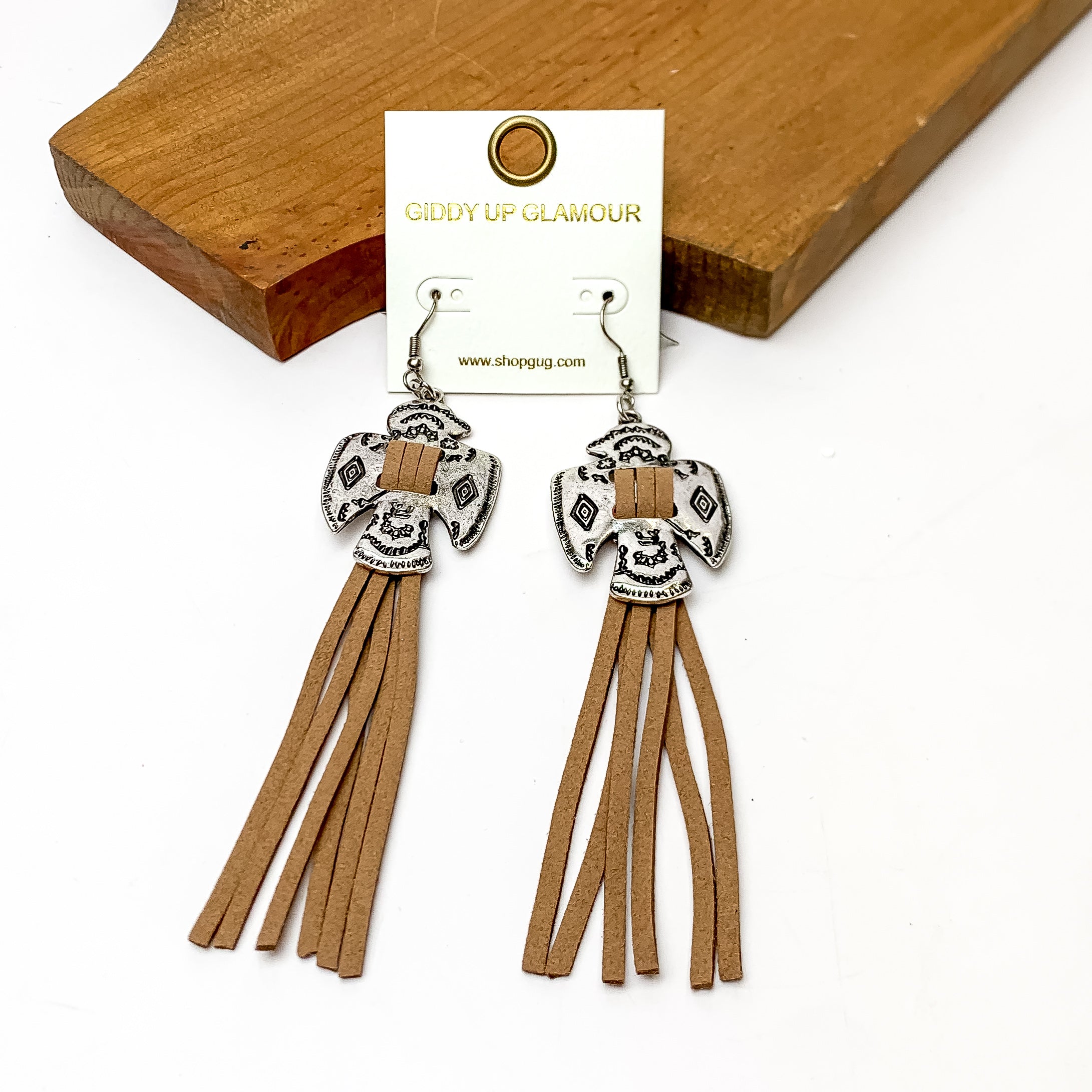 Thunderbird Tassel Earrings in Brown. These earrings are pictured laying against a wood piece. The background is white.