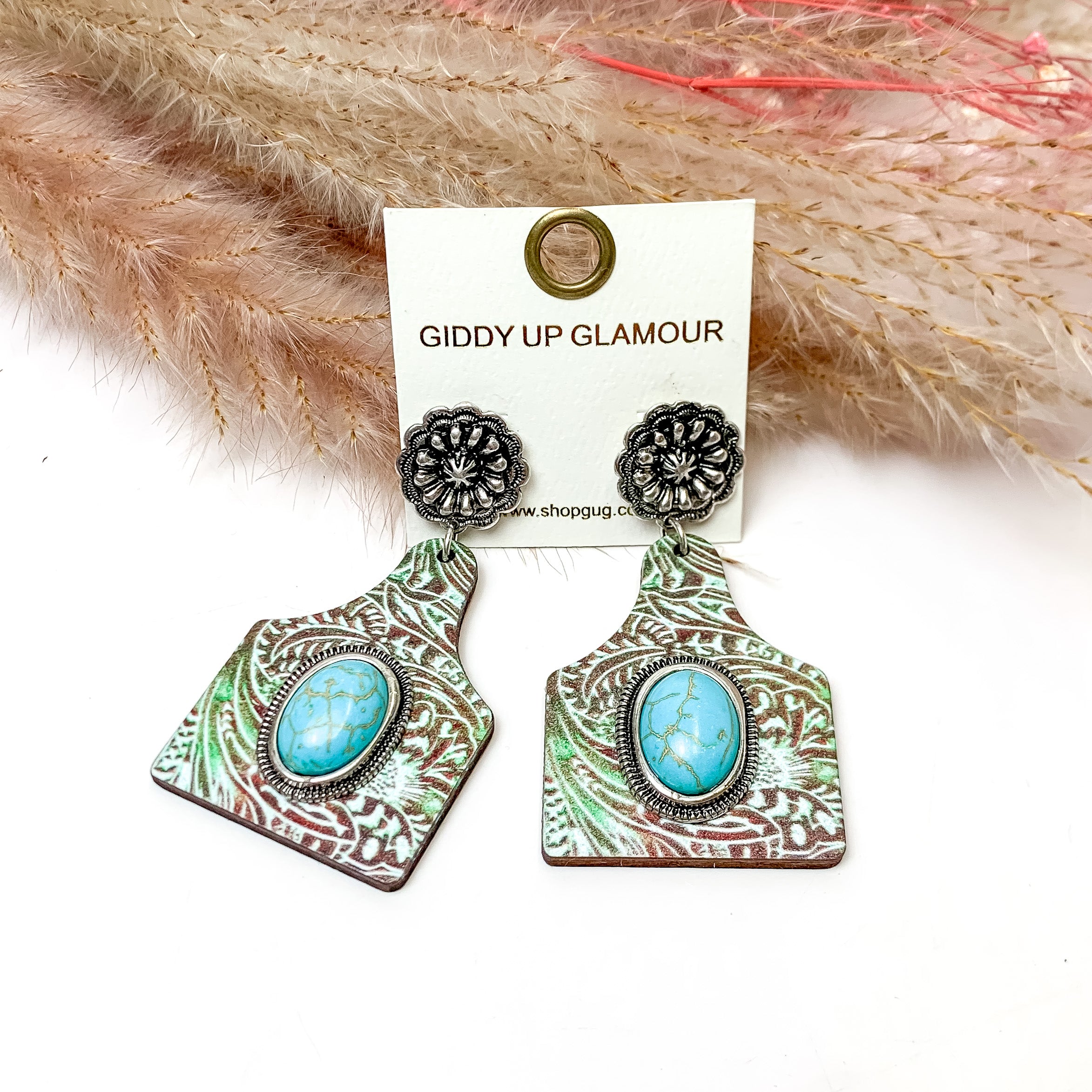 Patina Cattle Tag Earrings With Turquoise Center Stone. These earrings are pictured on a white background with flowers behind for decoration.