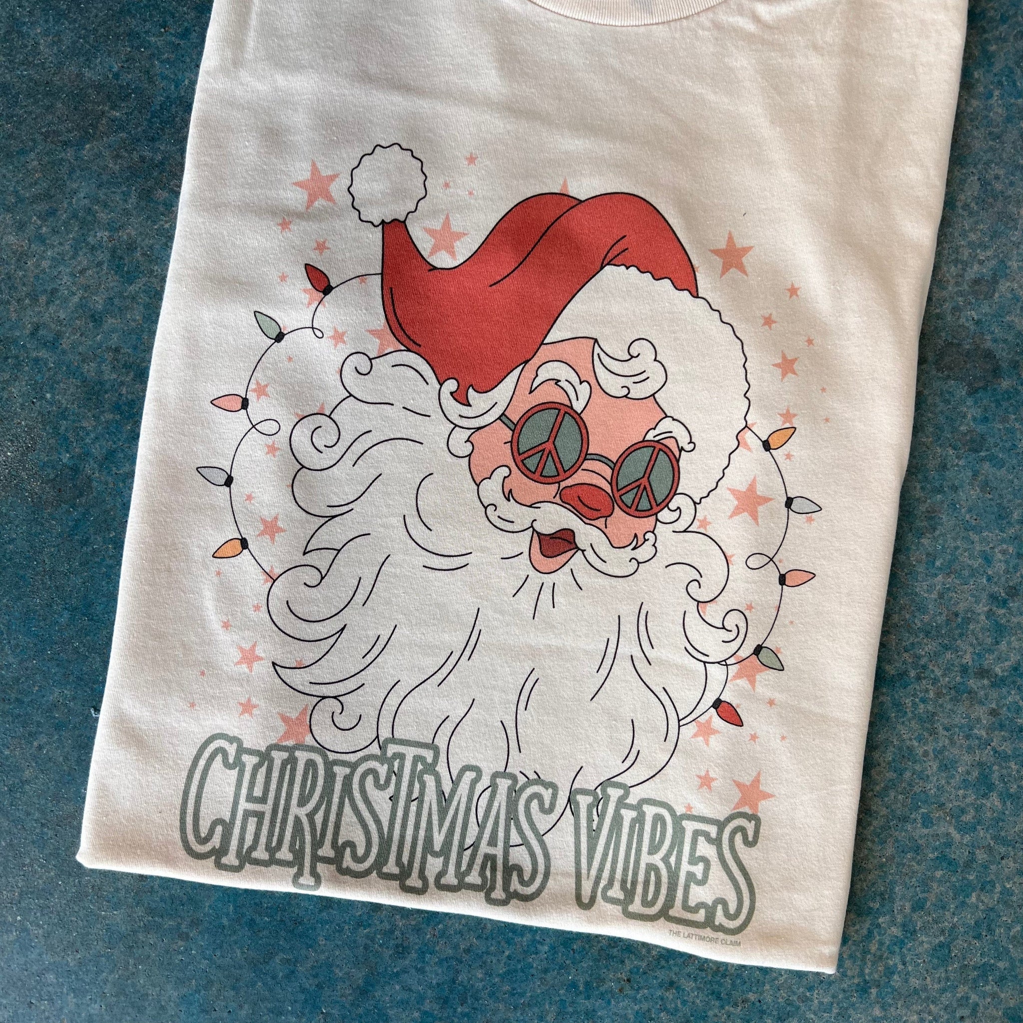 This crew neckline, short sleeve graphic tee is shown folded on a blue background. The graphic on this cream tee is a hand drawn Santa Clause wearing groovy peace sign glasses and the words "Christmas Vibes" are under his beard in a fun font outlined in green. There are also pink stars all around Santa in the background, as well as fun, bright colored string Christmas lights on either side of his beard. 