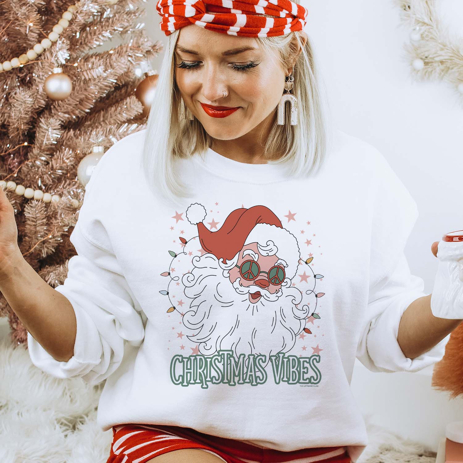 This white crew neckline graphic sweatshirt graphic has a hand drawn Santa Clause wearing a hat and groovy peace sign glasses. Pink stars of various sizes are all around him, as well as bright string lights on either side of his beard. At the bottom of his beard are the words "Christmas Vibes" in a fun font outlined in green. The model is sitting in front of a white background with a brown Christmas tree behind her right shoulder.
