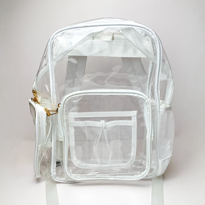 Pictured on a white background is a clear backpack with a white outline, white mesh pockets, and gold hardware. 