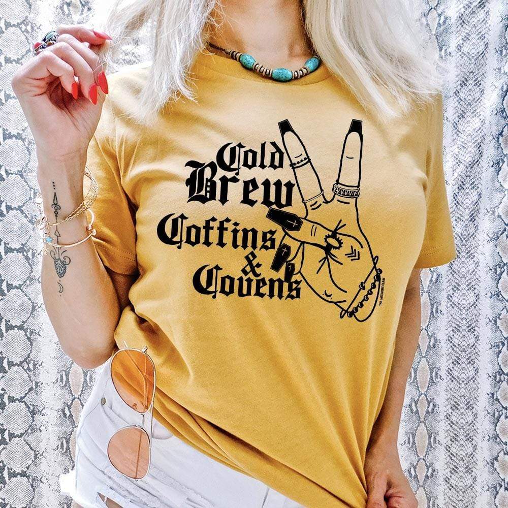 model is wearing a yellow graphic tee which reads "cold brew, coffins, and covens" in black font with a hand holding a peace sign. Model is standing in front of a snakeskin background. 