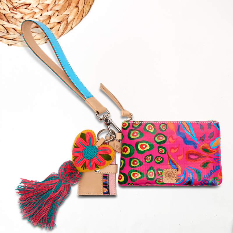 white background with wooden round decor in top left corner featuring the ceci consuela cloth fabric that is the sophie print in pink: pink background with paisley and fun swirls. the combi has a coin pouch, card slot organizer, fun charm and wristlet strap. perfect for on the go.