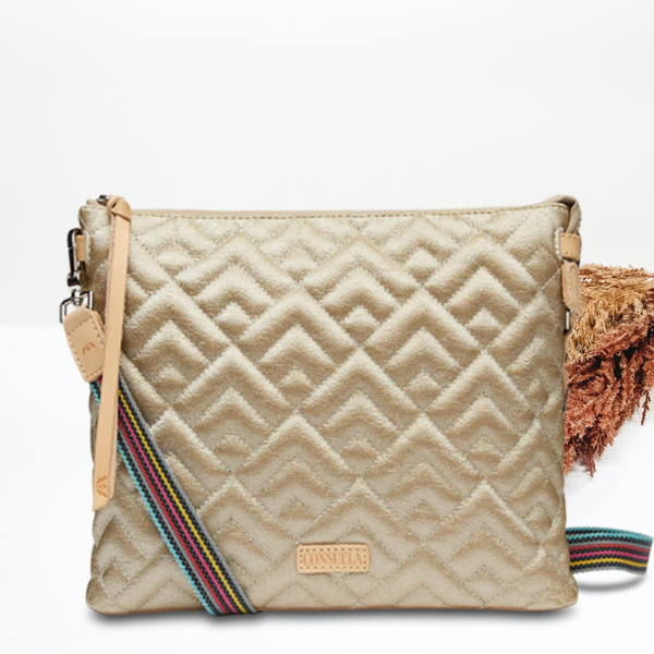 Pictured is a gold, diamond stitched downtown crossbody purse. This purse has a striped strap and a tan leather tassel on the zipper. This purse is pictured on a white background. 