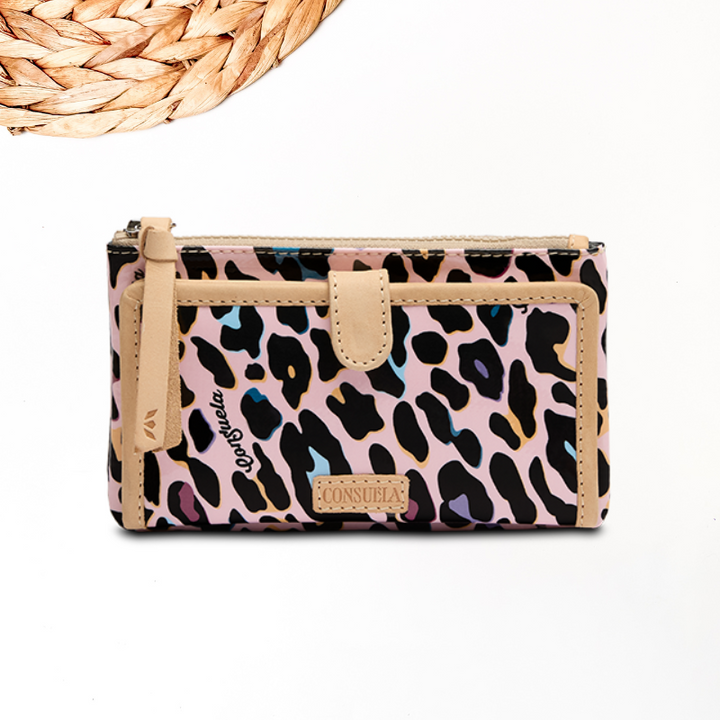 white background with wooden decor in the corner showcasing a pink leopard print consuela cloth wallet. this consuela cloth print is well known as deedee and recently coco and has been updated to ruth with a pink background and fun colors as the spots. this wallet features a zip closure for money, card slots and more.