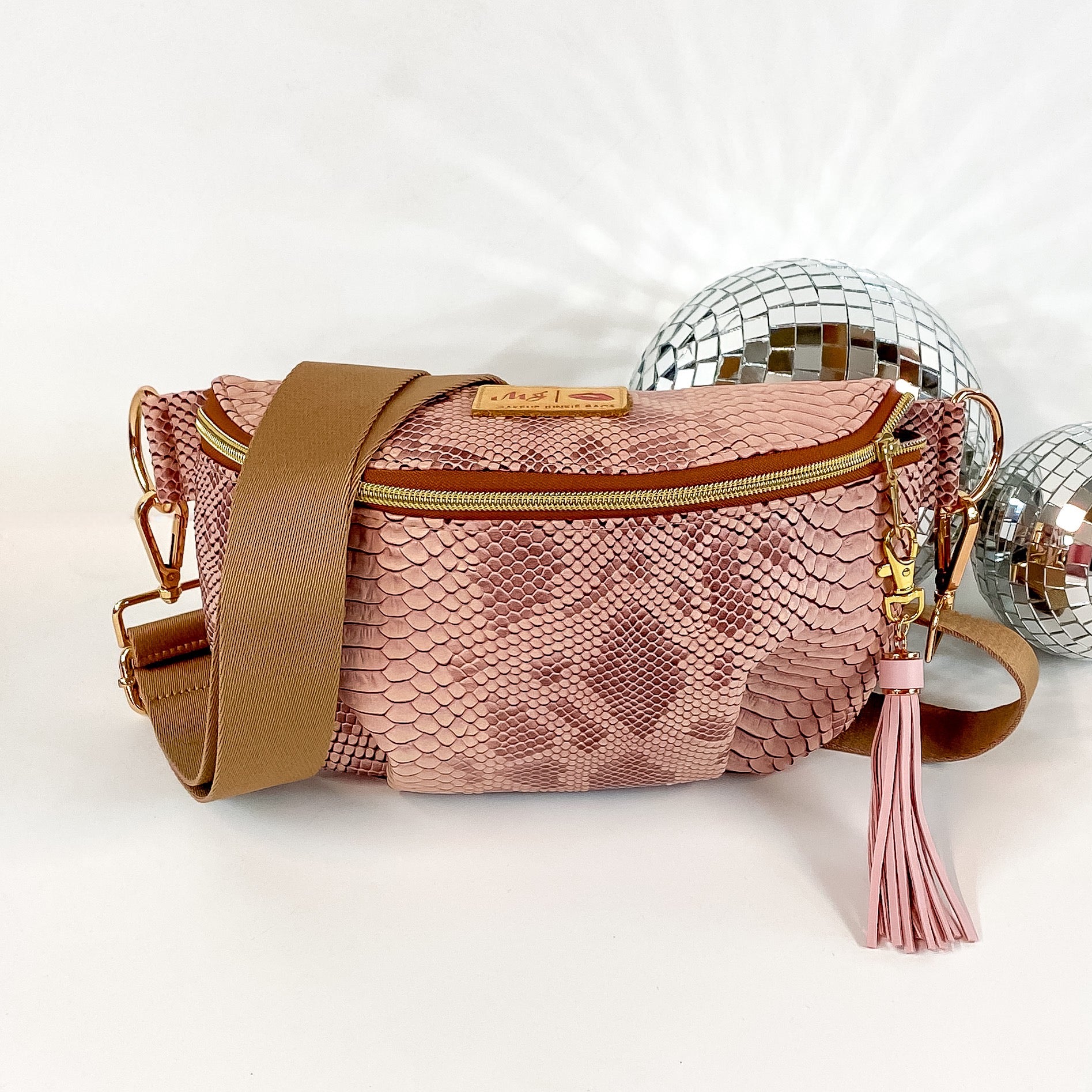 Pictured on a white background with disco balls in the background is a sidekick bag in a copper snake print. This bag includes a middle zipper and a tassel.