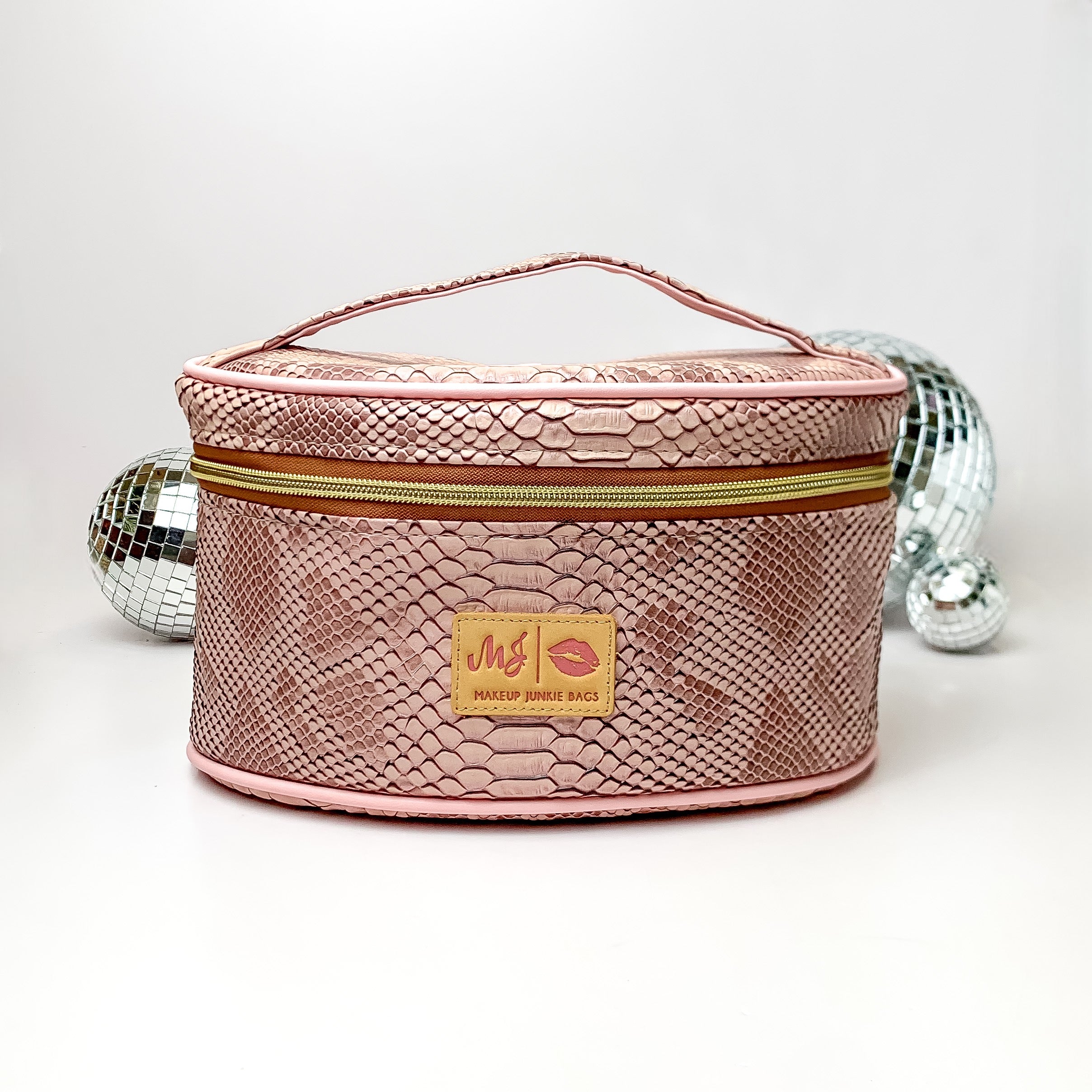Pictured on a white background with disco balls in the background is a traincase with a top handle in a copper snake print. This bag includes a middle zipper and a tassel.