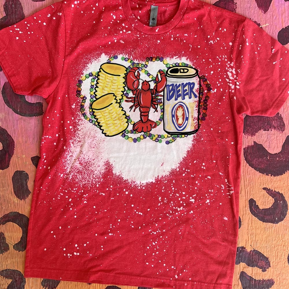 A red short sleeve tee with a bleach splatter pattern and a large bleach spot in the center featuring a graphic of a red crawfish with two corn on the cobs and a beer. Behind the crawfish are purple, green, and yellow mardi gras beads