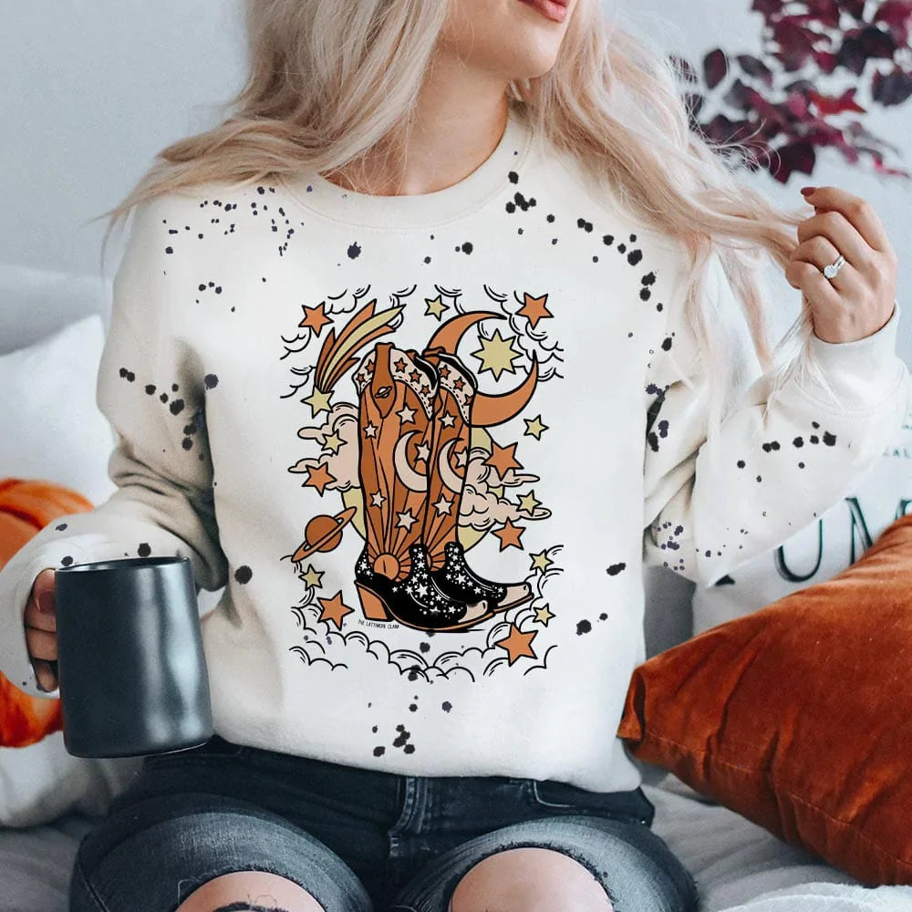 This white sweatshirt includes a crew neckline, long sleeves, and cute hand drawn design of brown and black cowboy boots with a cosmic scene surrounding them. The cosmic scene include a moon, planet, a shooting star, clouds, the moon, and other stars throughout in the cream rust orange, and pastel yellow colors. Black paint splatters are all over the sweatshirt as well. 