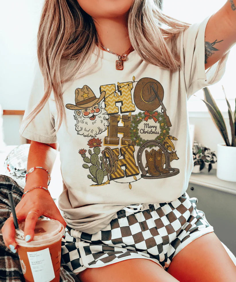 Cowboy Santa “Ho Ho Ho” graphic tee showing western Santa with hat, boots, wreath, and cacti on a cream tee.
