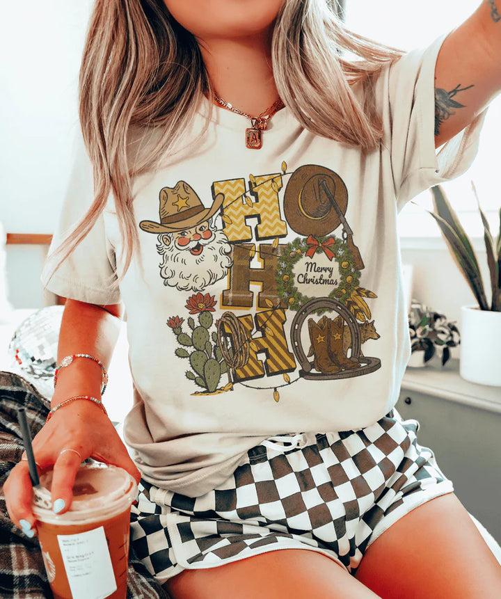 Cowboy Santa “Ho Ho Ho” graphic tee showing western Santa with hat, boots, wreath, and cacti on a cream tee.