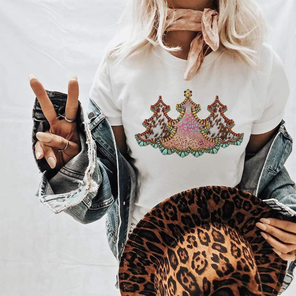 This white tee includes a crew neckline, short sleeves, and a cute Christmas graphic of three cowhide Christmas trees. The model has it styled here with a denim jacket, neutral wild rag, and leopard print felt hat. 