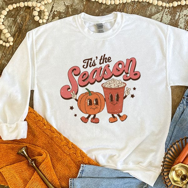 This white sweatshirt includes a crew neckline, long sleeves, and a cute hand drawn design of a pumpkin, coffee, and small stars with the words "Tis' the Season" above it. This graphic has fun Fall colors throughout including rust and orange. It is shown styled as a flat lay in this photo with the sleeves rolled and paired with light wash denim jeans.