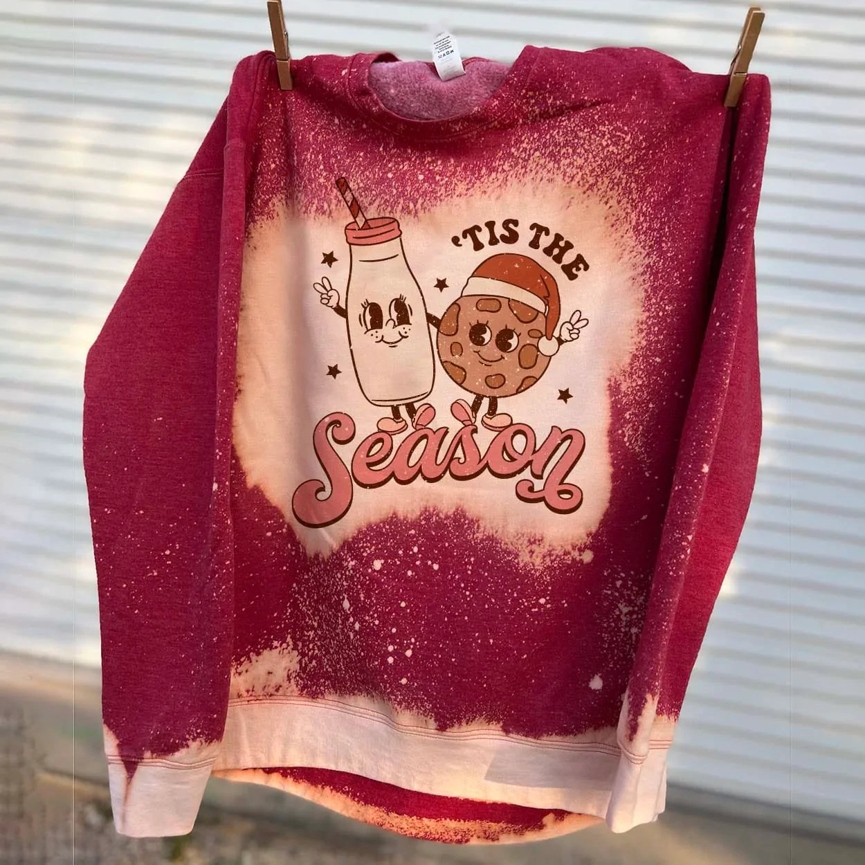 This bleached splatter sweatshirt in red includes a crew neckline, long sleeves, and a graphic that says "'Tis the Season" in a cute font with a cartoon milk and cookie wearing a Santa hat; both are throwing up peace signs with their outer hands and looking at each other. This sweatshirt is shown here hung from a clothesline with clothespins. 