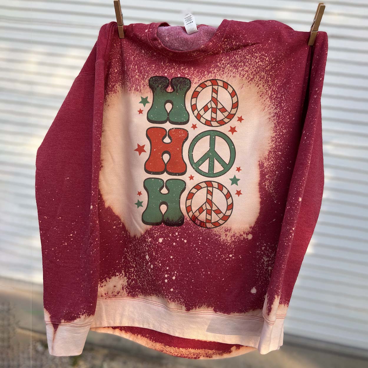 This bleached splatter sweatshirt in red includes a crew neckline, long sleeves, and a graphic that says "Ho Ho Ho" in a mix of cute font in red and green. The O's are peace signs and stars are all around the graphic. This is shown hanging from a clothesline with clothespins. 