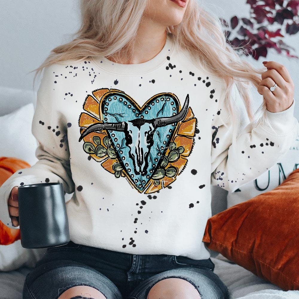 Model is wearing a white sweatshirt with black splatter paint all over. The sweatshirt has a bull skull graphic on a turquoise heart with yellow detailing and prickly pair cactus. Model has it paired with black distressed jeans.