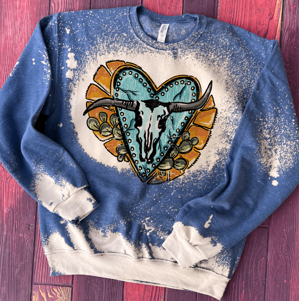 A blue long sleeve sweatshirt with bleach distressing. There is a bull skull graphic with turquoise heart, yellow detailing, and prickly pair cactus. Pictured on purple wooden background.