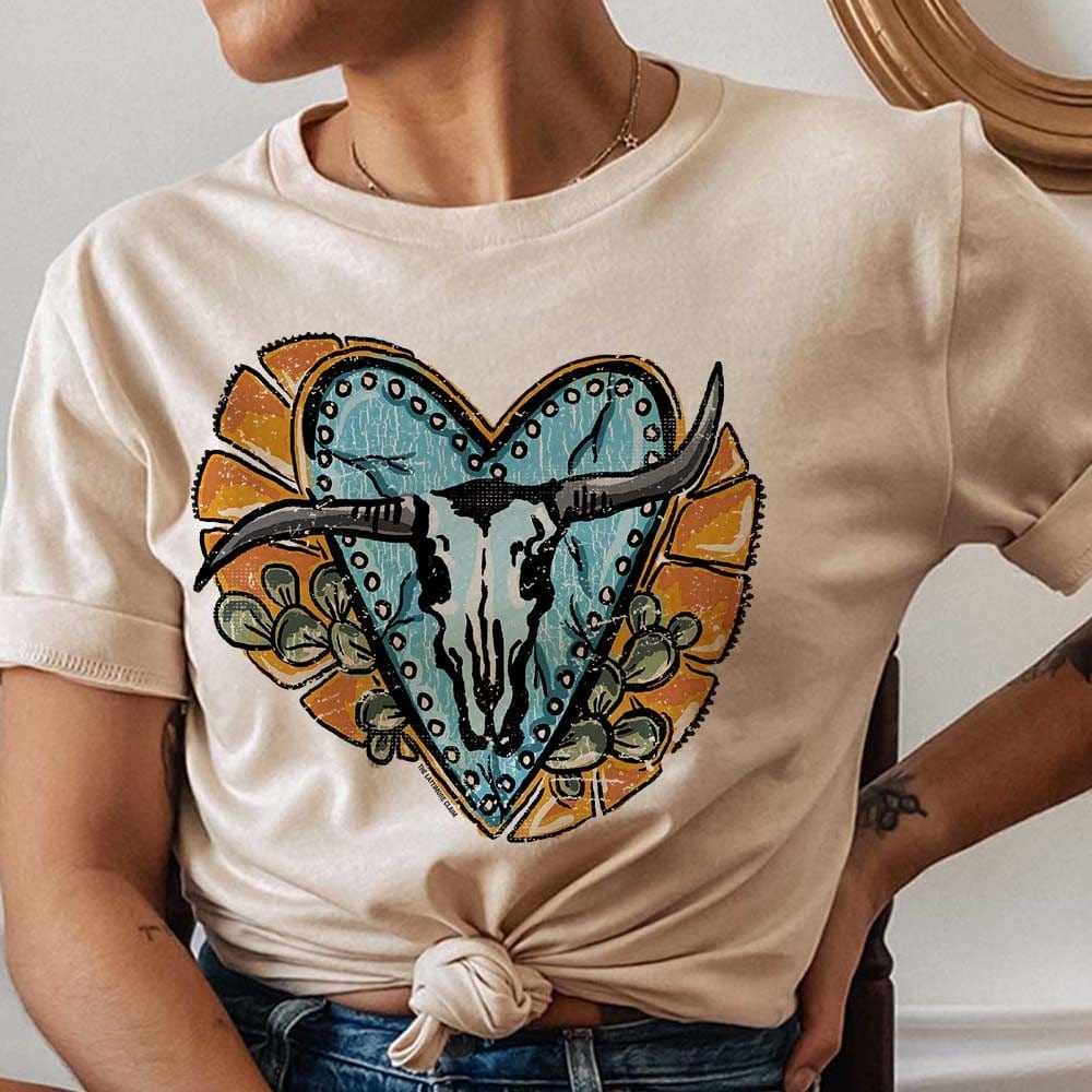 Model is wearing a cream tee with a graphic of a bull skull on a turquoise heart with yellow and cactus detailing. Model has the shirt knotted in the front.