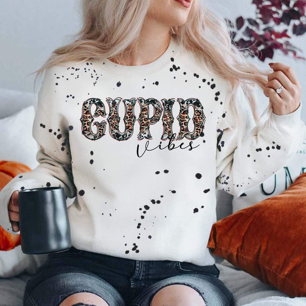 Model is wearing a cream colored and black splatter paint  sweatshirt shirt that has a front knot. The tee has a graphic that says "Cupid Vibes" in a leopard and cow print font.