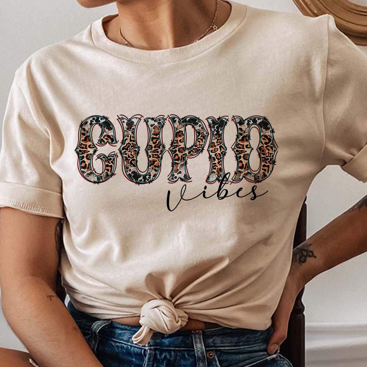 Model is wearing a cream colored tee shirt that has a front knot. The tee has a graphic that says "Cupid Vibes" in a leopard and cow print font.