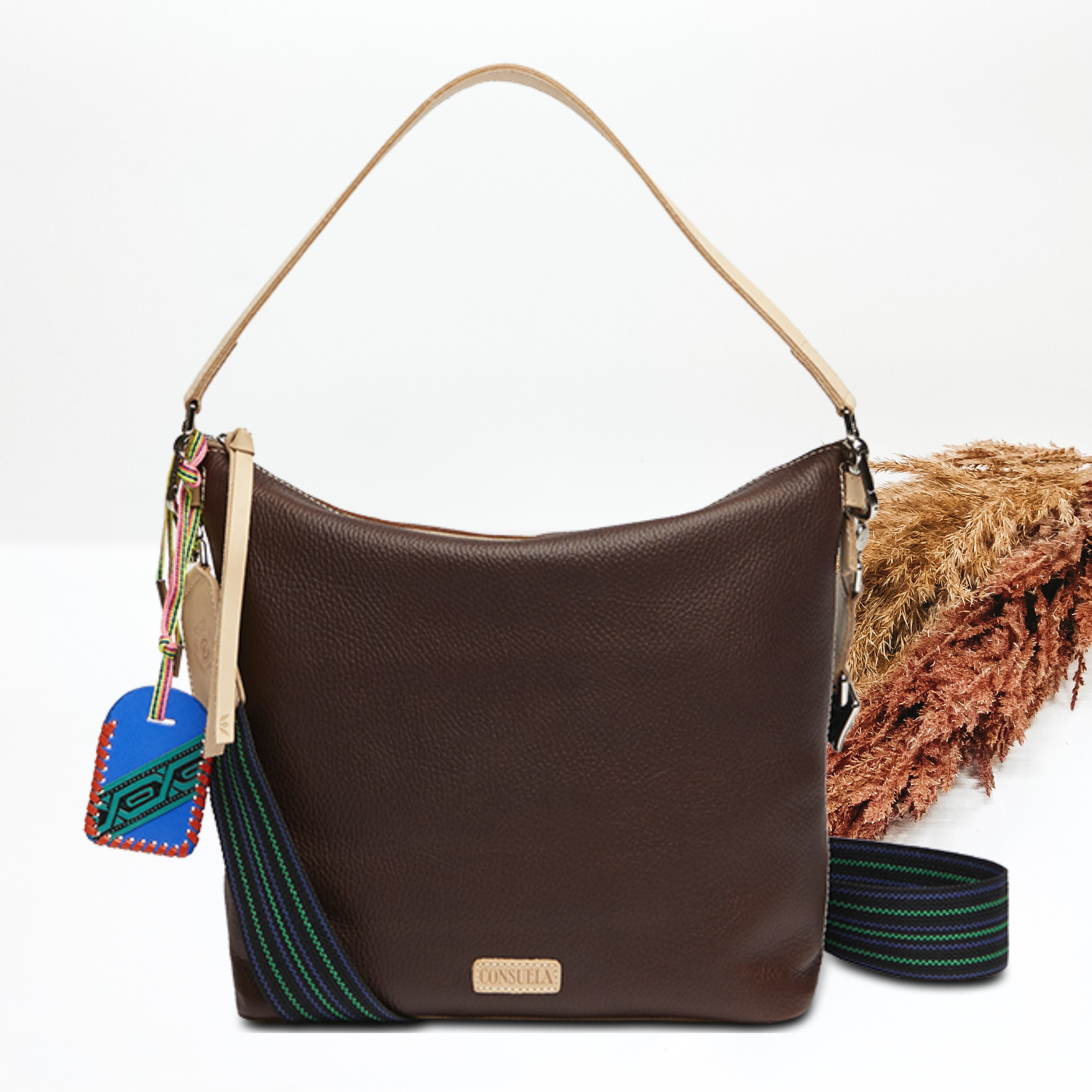 Pictured on a white background is dark brown leather hobo bag with a light tan shoulder strap. This bag also includes a striped crossbody strap., light tan tassel on the zipper and blue charm.