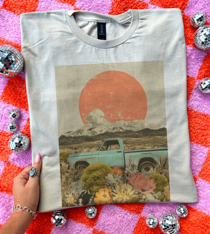 White t-shirt with a graphic of a truck and sunset held by a hand on a colorful surface with disco balls.