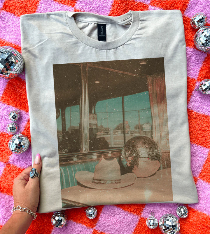 White t-shirt with a printed design of a room with hats and disco balls on a colorful background.