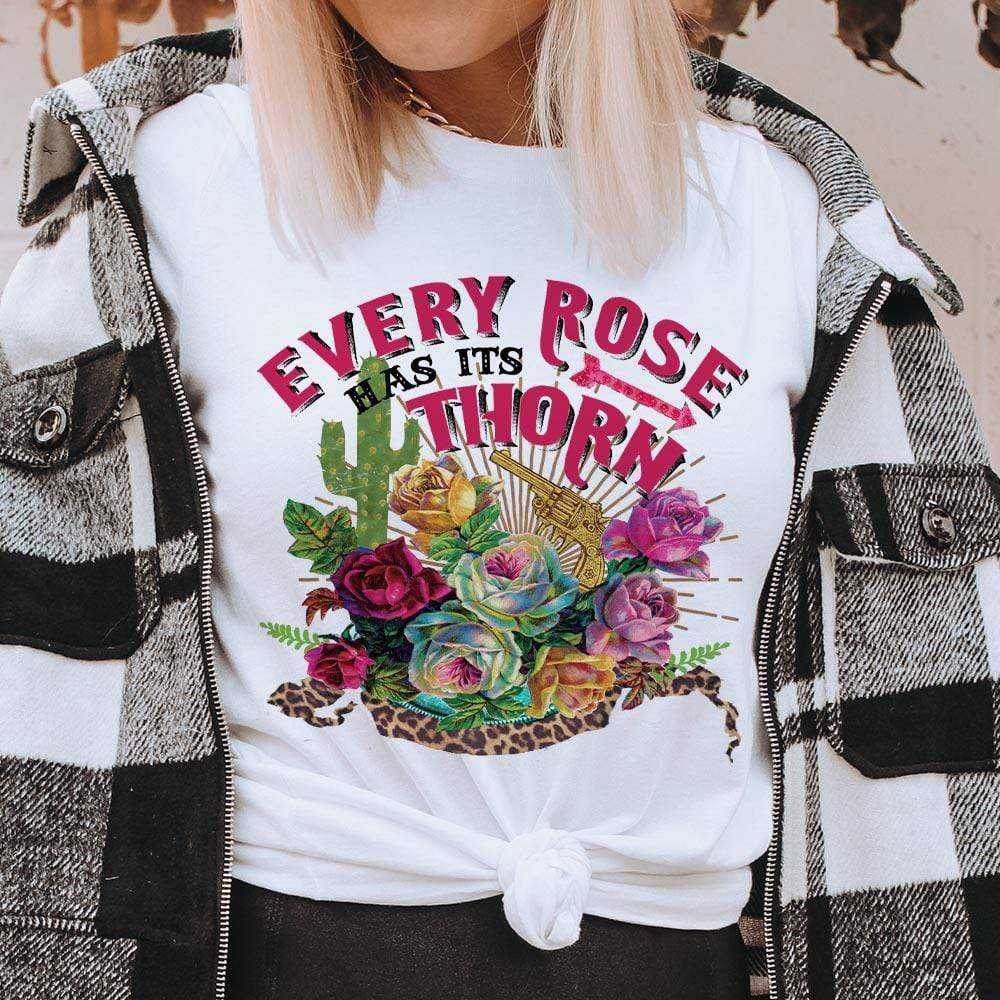 This graphic tee includes a crew neckline, short sleeves, and a bright, fun hand drawn design. The graphic on the tee is a mix of roses, cactus, a pistol, and the phrase "every rose has its thorn". This is being modeled with a thick chained necklace and a black and white plaid jacket. 