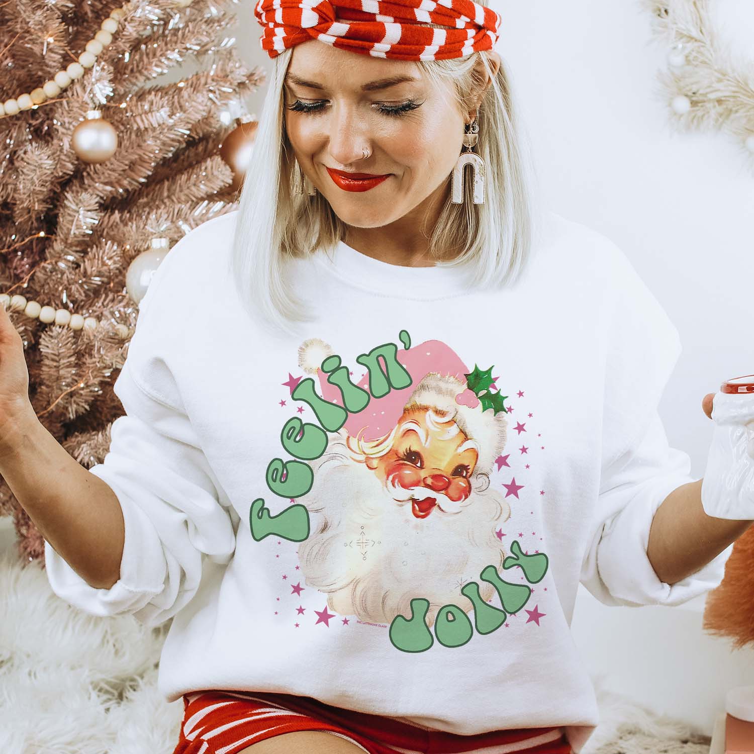 This white sweatshirt has a crew neckline, long sleeves, and a hand drawn graphic of a vintage Santa Clause, pink stars around him, and the words "Feelin' Jolly" in a fun, green bubble type of font. The Santa is also wearing a light pink Christmas hat. The tee is shown pictured here modeled with a pair of red and white striped shorts and matching headband, along with white dangle earrings. 