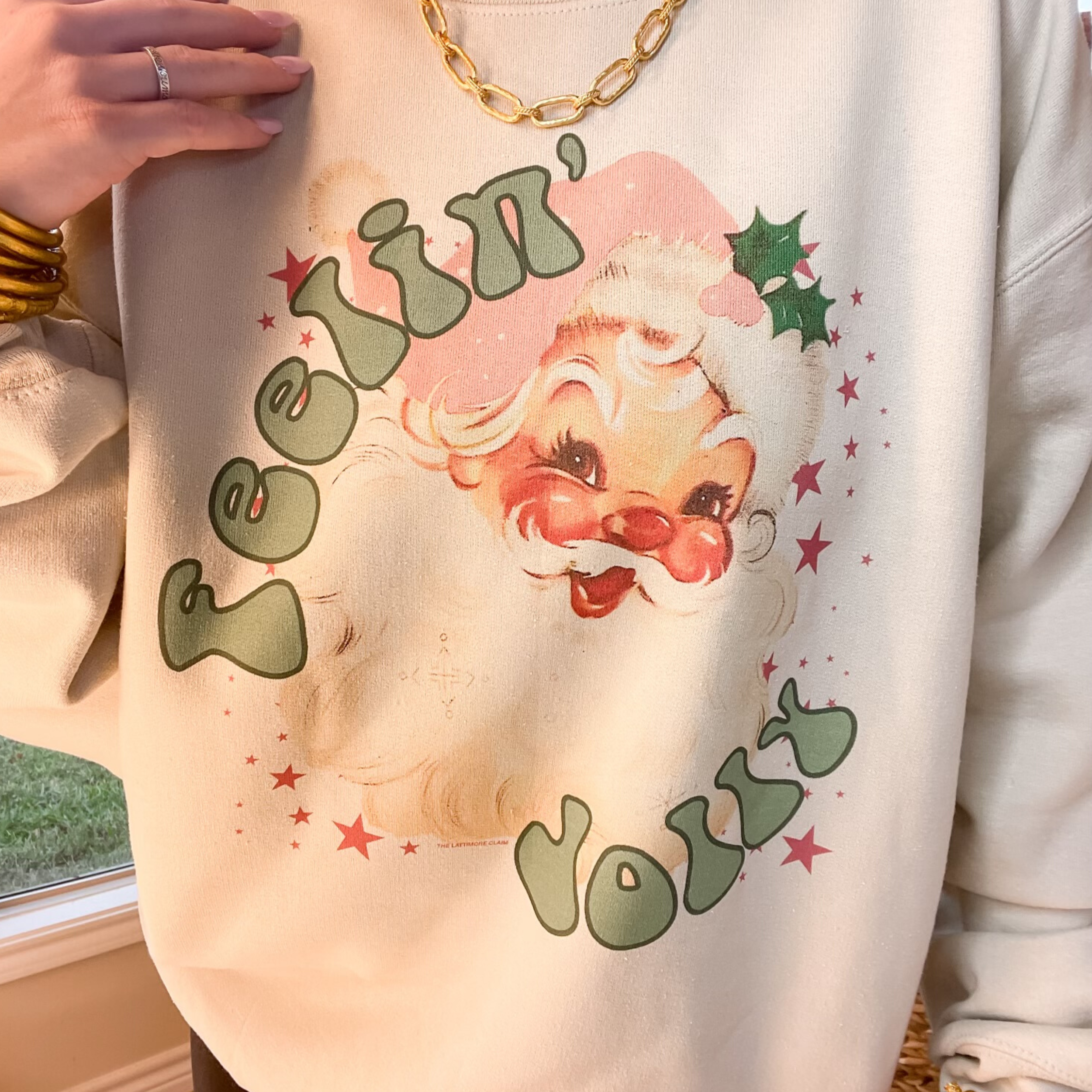 Photo features a cream sweatshirt with a graphic of santa and the words "Feelin' Jolly" in green. 