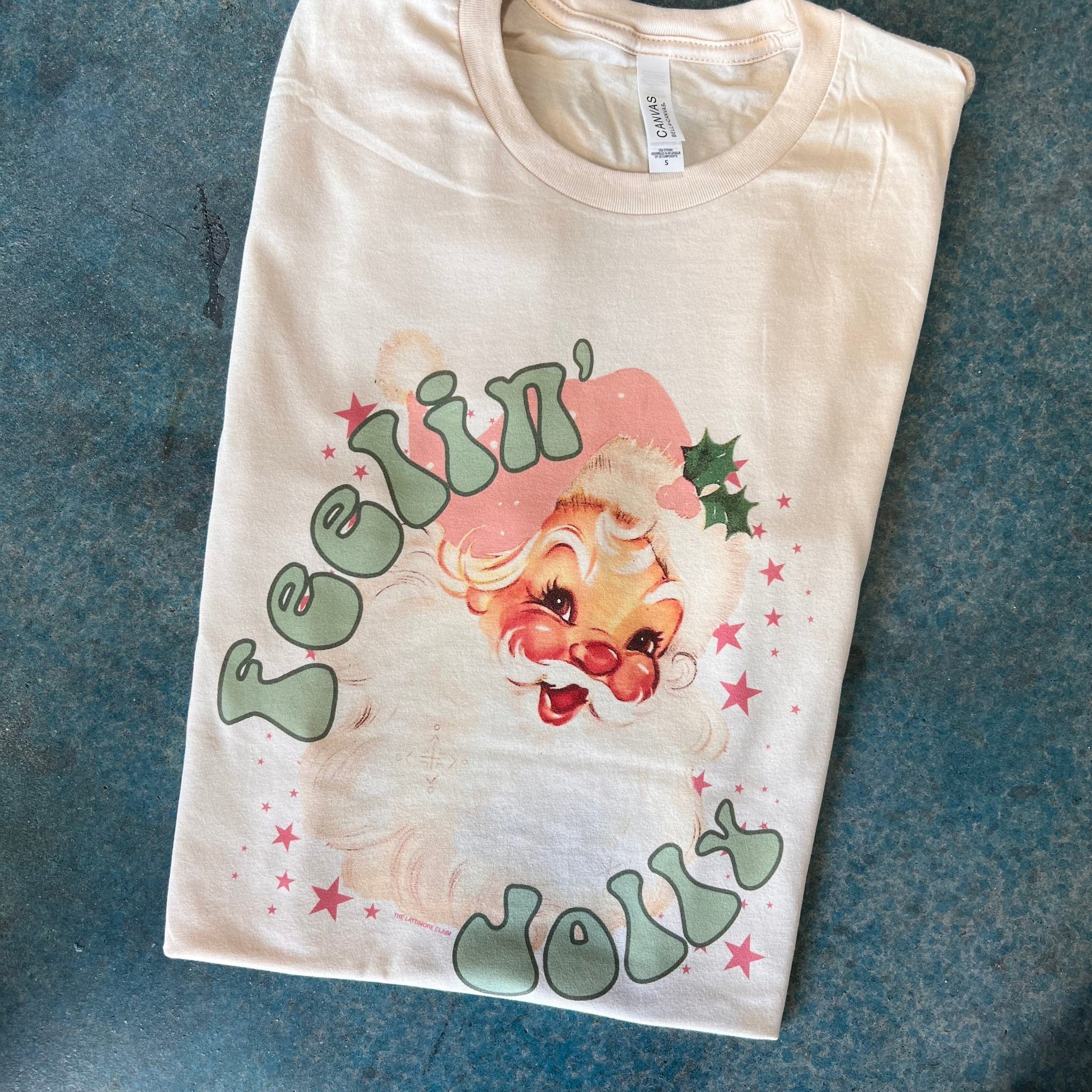 This cream tee includes a crew neckline, short sleeves, and a hand drawn graphic of a vintage Santa Clause, pink stars around him, and the words "Feelin' Jolly" in a fun, green bubble type of font.  The Santa is also wearing a light pink Christmas hat.  The tee is shown pictured here as a folded flatlay. 