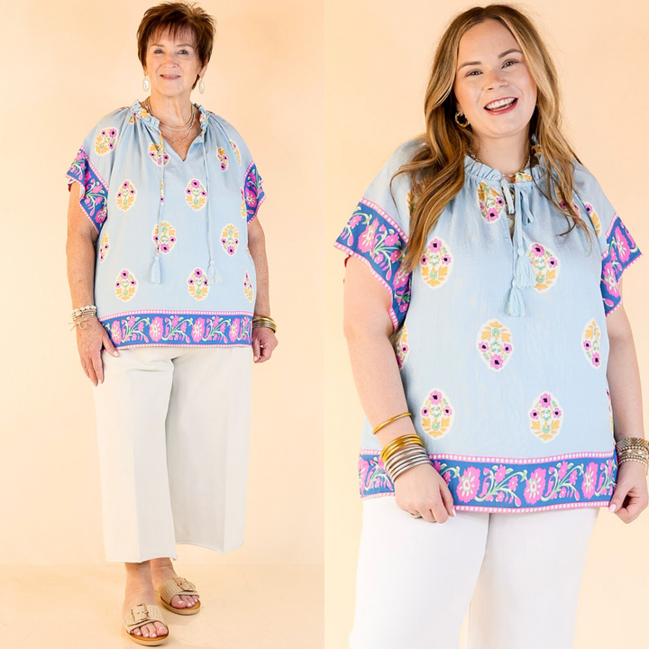 short sleeve A-line top with floral print details, a v neck with a tie closure, and is light blue with dark blue, pink, green, and yellow accent colors. Features a plus size model and the top comes in small through plus sizes.
