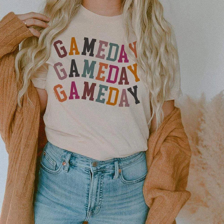 Model is wearing a cream colored short sleeve crewneck tee featuring a multicolor graphic of the words "Gameday"