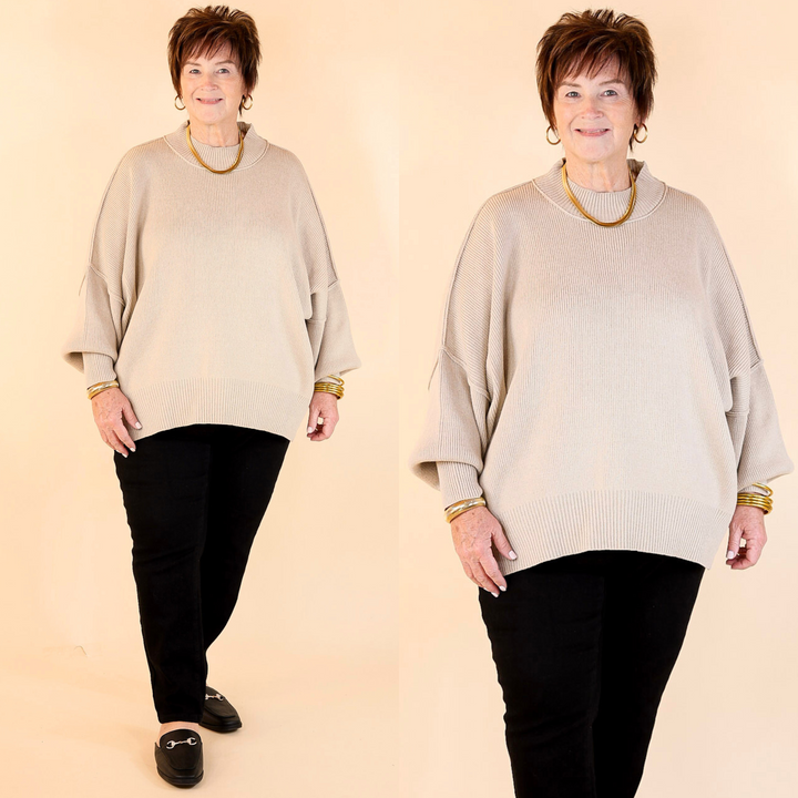 Model is wearing beige oversized sweater with side slit in combination with black pants and flat shoes. Against a plain background.