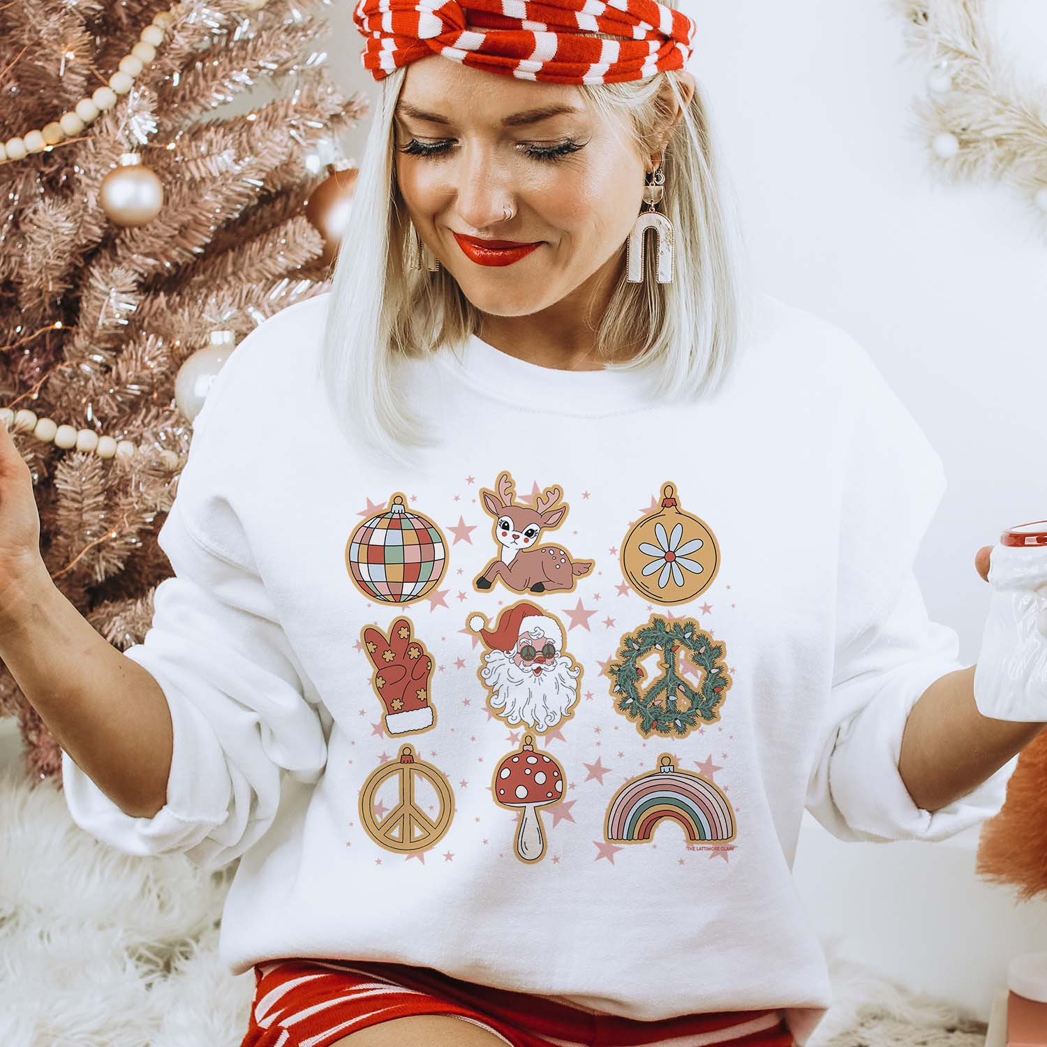 This white sweatshirt is being modeled with the sleeves pushed up and a red and white striped headband and matching shorts. The sweatshirt has 9 groovy and festive hand drawn Christmas artwork, such as a disco ball, reindeer, ornament, Santa Clause, and more! The background behind the model is white and a brown Christmas tree is over her right shoulder. She is also wearing white dangle earrings to complete the look. 