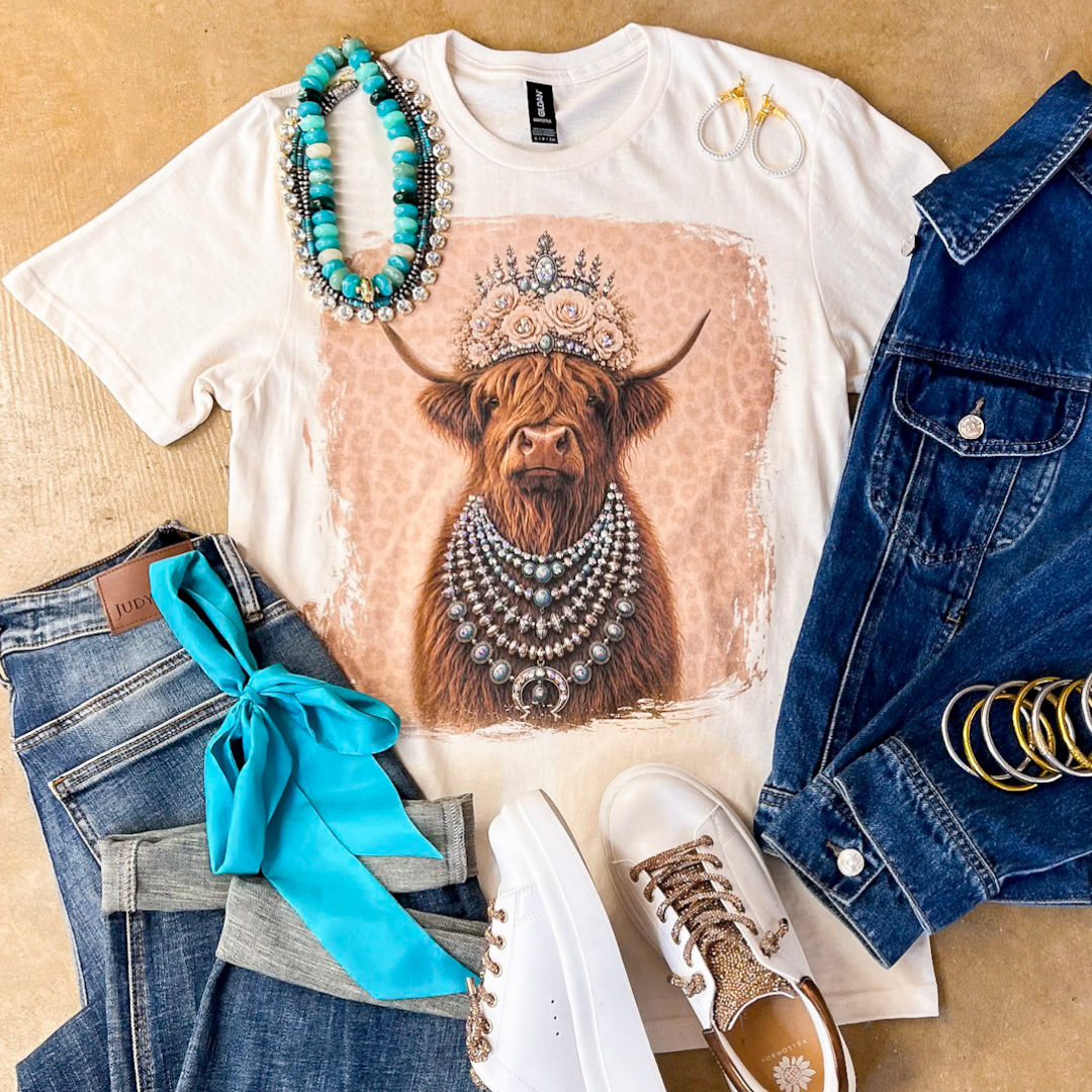 Heidi the Highland Queen crystal graphic tee in cream with a highland cow wearing a jeweled floral crown and layered necklaces on a pink leopard background, styled with denim and turquoise jewelry