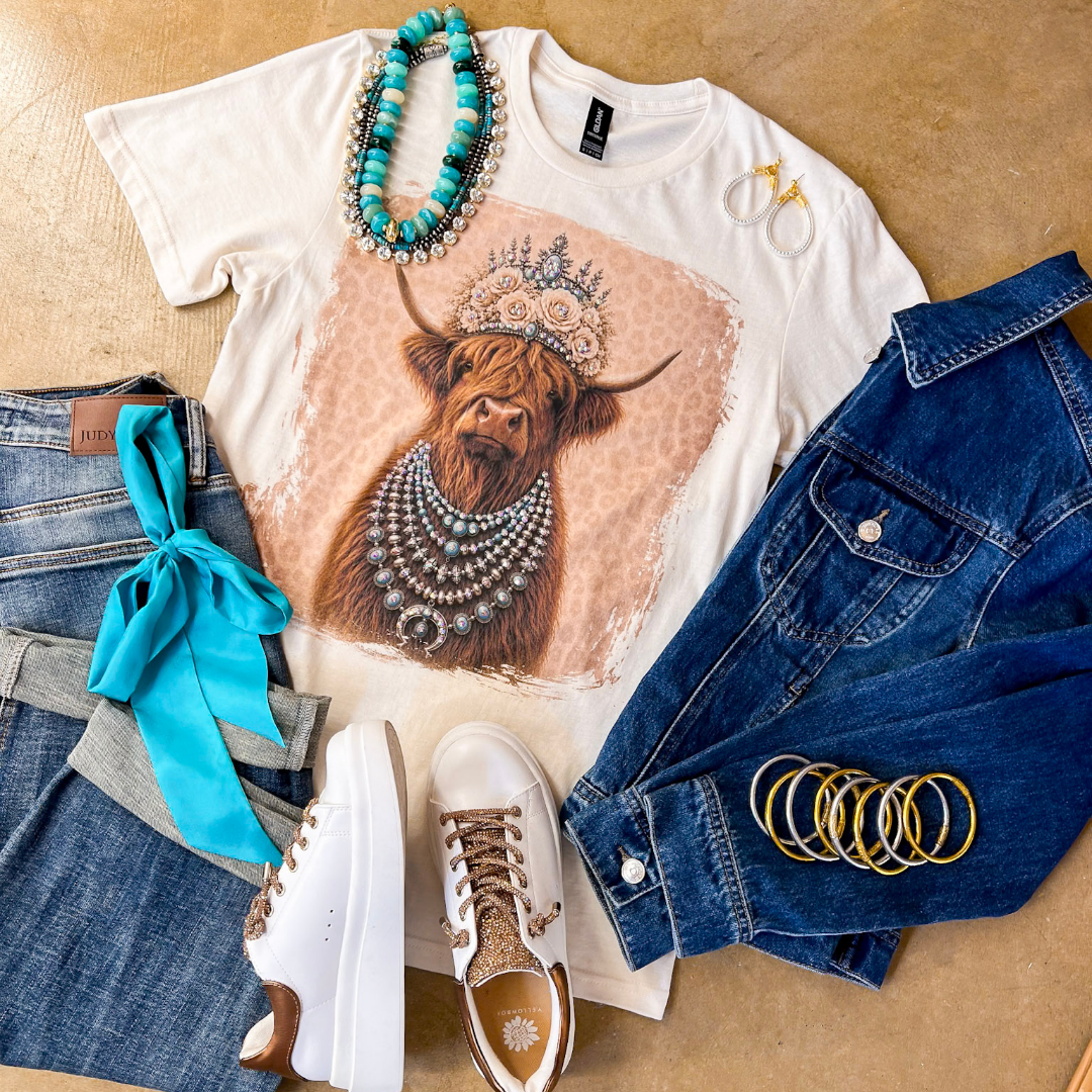 Heidi the Highland Queen crystal graphic tee in cream with a highland cow wearing a jeweled floral crown and layered necklaces on a pink leopard background, styled with denim and turquoise jewelry